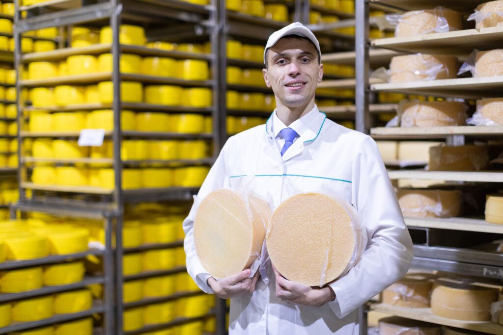 Un fromager fier. Ce dernier représente l'essence de l'artisanat et de la passion qui vont dans chaque meule de Comté. Source: <a href="https://www.freepik.com/free-photo/technologist-white-robe-with-yellow-cheese-head-his-hands-is-shop-production-butter-cheese-production-process-plant-dairy-products-racks-with-cheese_26151124.htm#query=fromager%20avec%20tablier&position=2&from_view=search&track=ais">Image by usertrmk</a> on Freepik